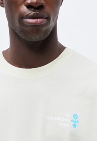 Κλείσιμο σε άτομο που φοράει λευκό πουκάμισο με το κείμενο «All Conditions Gear» και μια μικρή μπλε ψηφιακή σχεδίαση λουλουδιού στο στήθος.