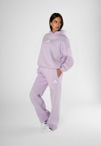 Felpa con cappuccio lavanda e pantaloni jogger abbinati, realizzati in morbido materiale. Presenta un logo bianco sul petto e sulla coscia superiore. Sneakers bianche.