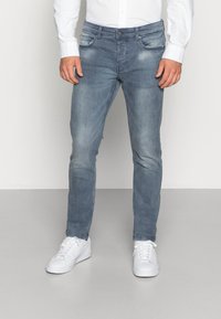 Vaqueros de mezclilla azul claro con un corte slim, diseño de cinco bolsillos y detalles deslavados. Combinados con zapatillas blancas y una camisa blanca de botones.