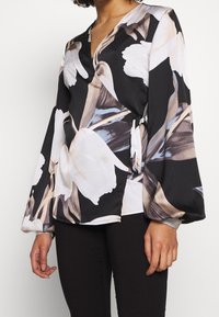 Blouse enroulée noire à motifs floraux avec de grandes fleurs blanches et beiges, décolleté en V profond, longues manches évasées et détail de taille nouée.