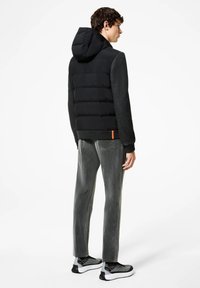 Gilet imbottito nero con cappuccio, maniche grigie testurizzate e accentuazione arancione. Abbinato a pantaloni grigi sbiaditi e sneaker nere e grigie.