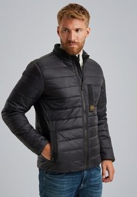 Chaqueta acolchada negra con cuello alto, cierre de cremallera y bolsillos laterales. Presenta costuras horizontales y un parche con el logotipo en el pecho.