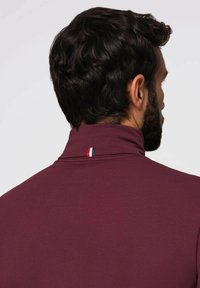 Burgundy polo skjorta med hög krage, med en liten blå, vit och röd detalj på baksidan. Slät tyg och figurnära design.
