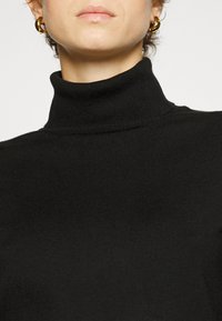 Un col roulé noir en tricot côtelé, avec un col haut et un design sans couture autour du cou. Boucles d'oreilles en or en forme de créole visibles.