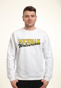 Bílý svetr s nápisem "MICHIGAN" velkými žlutými písmeny a "Wolverines" černým písmem, kulatým výstřihem a dlouhými rukávy.