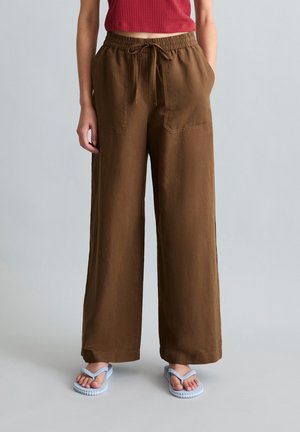 Femme portant un pantalon marron à jambes larges avec poches avant et taille à cordon, associé à des tongs bleues sur fond gris.