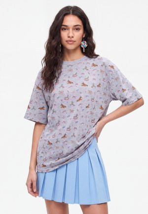 Ung kvinde med langt mørkt hår iført en grå mønstret T-shirt, blå plisseret nederdel og farverige øredobber formet som slikkepinde, står med hånden på hoften.
