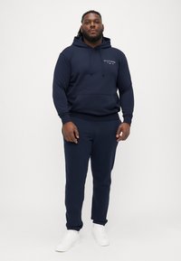 Ναυτικό μπλε φούτερ με μπροστινή τσέπη και κουκούλα με κορδόνι, συνδυασμένο με αντίστοιχους joggers. Λευκά αθλητικά παπούτσια ολοκληρώνουν το σύνολο.