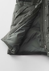 Grüne Pufferjacke mit Reißverschluss, elastischen Bündchen und gepolstertem Innenfutter, das eine glatte, matte Oberfläche und ein Logo-Detail aufweist.