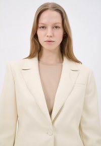 Blazer color crema con un design strutturato, rever a punta, chiusura con un singolo bottone e un sottile dettaglio della tasca frontale, indossato sopra un top beige.
