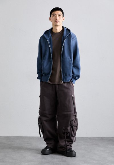Blå zip-up hettegensere over en brun crew neck skjorte, kombinert med mørke cargo bukser med glidelåser og lommer, samt svarte joggesko.