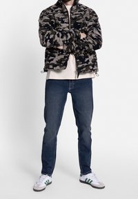 Bunda typu bomber z fleecu v maskáčovém vzoru, šedá a námořnická modř. V kombinaci s modrými džínami a bílými teniskami se zelenými akcenty. Minimalistický design.
