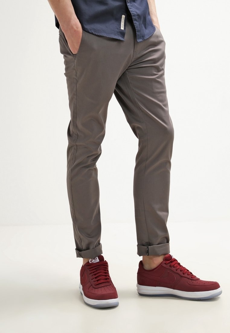 Personne portant un pantalon slim gris roulé aux chevilles et des baskets rouges à semelles blanches, debout les mains dans les poches.