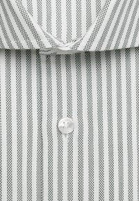 Gros plan d'une chemise blanche avec des rayures verticales noires en pointillés et un seul bouton blanc sous le col.