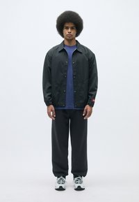 Svart lättviktig jacka med tryckknappar över en blå skjorta, kombinerad med mörka sweatpants och vita sneakers, framför en neutral bakgrund.