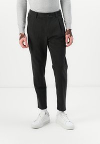 Pantalons noirs fuselés avec une taille élastique et des plis à l'avant. Portés avec des baskets blanches et un haut à manches longues gris clair. Tissu doux.