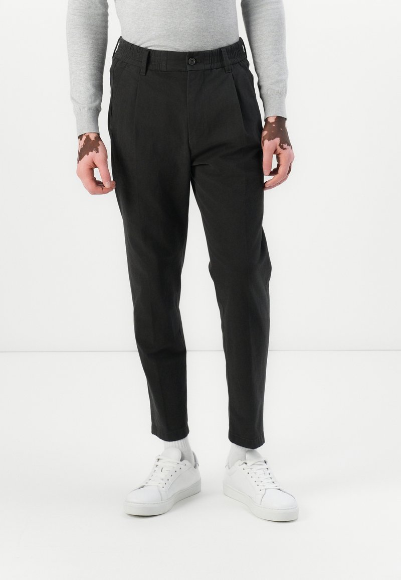 Pantalons noirs fuselés avec une taille élastique et des plis à l'avant. Portés avec des baskets blanches et un haut à manches longues gris clair. Tissu doux.