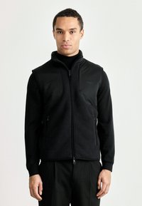 Colete preto com fecho à frente, feito de fleece macio, apresentando um colarinho alto, dois bolsos laterais com fecho e um pequeno logotipo no peito. Textura suave e corte moderno.