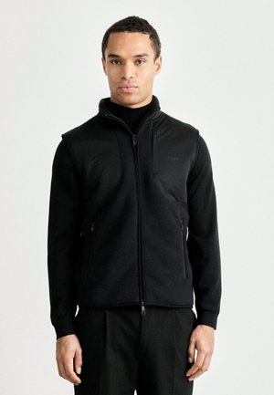 Gilet nero con zip realizzato in morbido pile, dotato di un collo alto, due tasche laterali con zip e un piccolo logo sul petto. Texture liscia e vestibilità moderna.