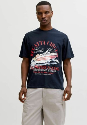 Muž v čiernom tričku s červenou a bielou grafikou jachtárskych pretekov a svetlobéžovými nohavicami na neutrálnom pozadí.