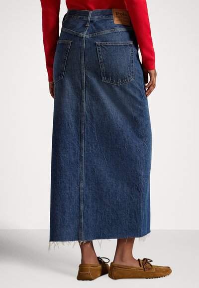 Žena oblečená v dlhej modrej rifľovej sukni s vreckami vzadu, červenom svetri a hnedých mokasínach stojaca na jednoliatom pozadí.