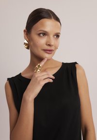 Złote kolczyki w kształcie obręczy z teksturowanym wzorem oraz abstrakcyjnym pierścieniem w polerowanym wykończeniu, noszone z czarną bezrękawnikową bluzką.