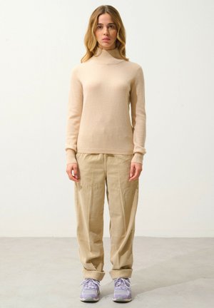 Jersey beige de cuello alto en un material de punto suave combinado con pantalones de pana claros. El modelo lleva zapatillas deportivas moradas con cordones claros.
