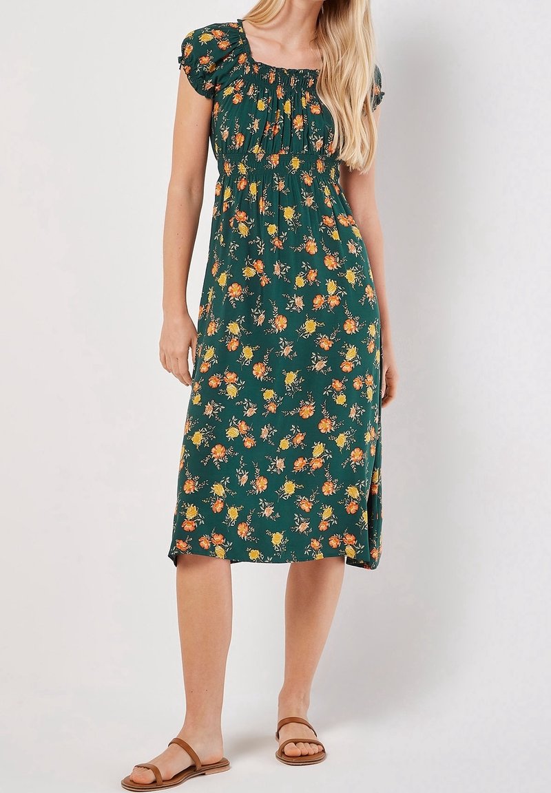 Femme portant une robe midi verte imprimée de fleurs orange et jaune, corsage froncé, manches bouffantes et sandales plates marron.
