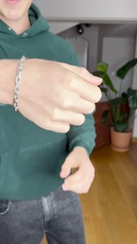 Železna verižica na zapestju. Verižica vsebuje prepletene člane z polirano površino. Vidna je zelena majica.