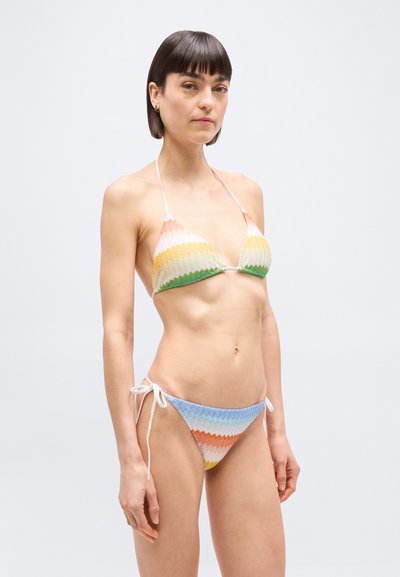 Γυναίκα που φοράει πολύχρωμο μπικίνι με ζ ιγκ-ζαγκ σχέδιο, τοπ τριγωνικό και παντελόνι με κορδόνια στο πλάι, στέκεται μπροστά σε απλό λευκό φόντο.