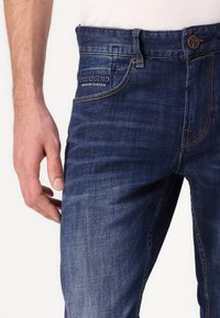 Mörkblå denimjeans med slim fit, featuring en synlig logopatch på midjan, kontrasterande sömmar och klassisk femfickdesign.