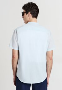 Uomo che indossa una camicia a maniche corte azzurro chiaro e pantaloni scuri, girato di spalle, con occhiali da sole neri e capelli corti ricci.