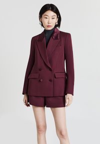 Blazer bordeaux doppiopetto con rever in raso e due tasche laterali, abbinato a pantaloni corti coordinati. Texture liscia, vestibilità su misura.