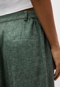 Záber zblízka na osobu oblečenú v zelených textúrovaných nohaviciach s vreckom na chrbte a elastickým pásom, v kombinácii s bielym rebrovaným topom.