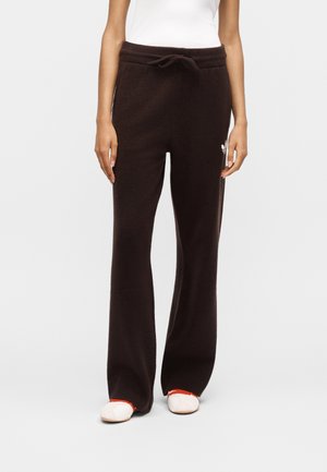 Donna che indossa pantaloni neri con coulisse, piccolo logo bianco sulla coscia sinistra e scarpe bianche con dettagli rossi.