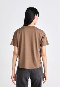 Camiseta de algodón marrón con un corte relajado, mangas cortas y un escote redondeado. La tela parece suave y ligera. Llevada con jeans de color gris oscuro.