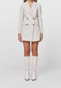 Biała marynarka w kształcie sukienki z podwójnymi złotymi guzikami, klapami w serek i dwoma przednimi kieszeniami; zestawiona z białymi botkami do kolan.