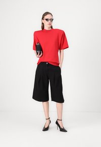 Rode oversized top met korte mouwen, zwarte geplooide shorts en zwarte hoge hakken. Model houdt een kleine zwarte tas vast en draagt een zonnebril.