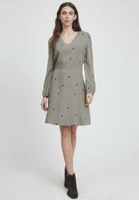 Grünes Kleid mit schwarzen Pünktchen, V-Ausschnitt, langen Ärmeln, tailliertem Schnitt, ausgestellt Rock. Der Stoff wirkt glatt mit einem leichten Glanz.