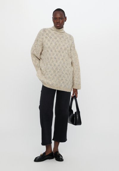 Beige gebreide trui met een hoge halslijn, voorzien van een ruitpatroon. Gecombineerd met zwarte rechte jeans en zwarte instappers.