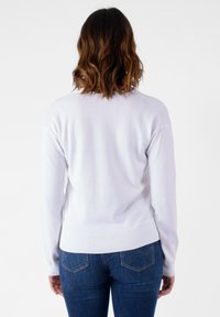 Maglione bianco lavorato a maglia con maniche lunghe, polsini e orlo a costine, vestibilità rilassata. Indossato con jeans blu, che mettono in risalto la texture liscia e la semplicità.
