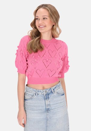 Femeie zâmbitoare cu păr ondulat poartă un pulover roz scurt, cu modele în formă de inimă și ciucuri, asortat cu o fustă din denim bleu deschis.