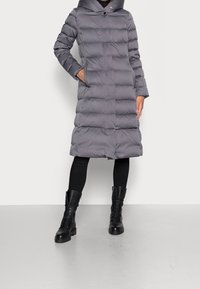 Abrigo largo acolchado gris con capucha, cierre de botones y bolsillos laterales. Se lleva con mallas negras y botas altas con cordones hasta la rodilla.