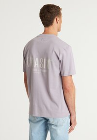 T-shirt a maniche corte color lilla con grande testo bianco stampato sul retro, in morbido cotone e design a vestibilità rilassata.