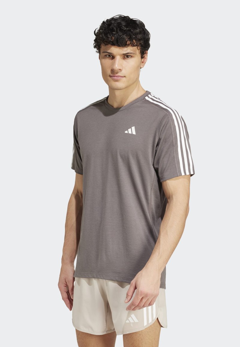 Camiseta deportiva gris de manga corta, con cuello redondo y detalles en blanco de tres rayas en los hombros. Presenta un pequeño logo blanco en el pecho.