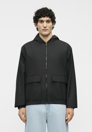 Νέος άνδρας που φοράει μαύρο ζακέτα με φερμουάρ και κουκούλα, με εμπρόσθιες τσέπες με καπάκι και ανοιχτό μπλε τζην, στέκεται μπροστά σε απλό φόντο.