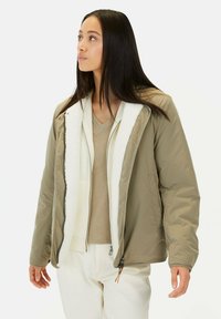 Khakifarbene, isolierte Jacke mit Kapuze, ausgestattet mit einem fleecegefütterten Kragen und einem Reißverschluss vorne. Getragen über einem beigefarbenen V-Ausschnitt-Oberteil, kombiniert mit weißen Hosen.