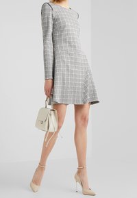 Mujer con un vestido de cuadros en blanco y negro de manga larga, sosteniendo un bolso beige, usando zapatos de tacón beige, de pie frente a un fondo blanco.