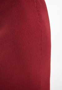 Geribbeld rood stof met verticale strepen; close-up weergave benadrukt de textuur en stikdetails langs de zijnaad.