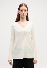 Sweter z dekoltem w serek, z długim rękawem w kolorze ecru, o fakturze prążkowanej i luźnym kroju, z nieco szerszym prążkowaniem na całej powierzchni.